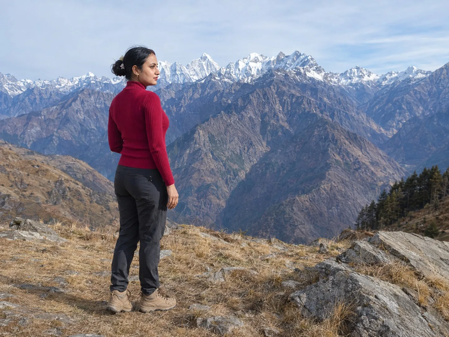 Vandana on dry grass at a ridge edge looking out over brown mountainsides towards jagged snow peaks.