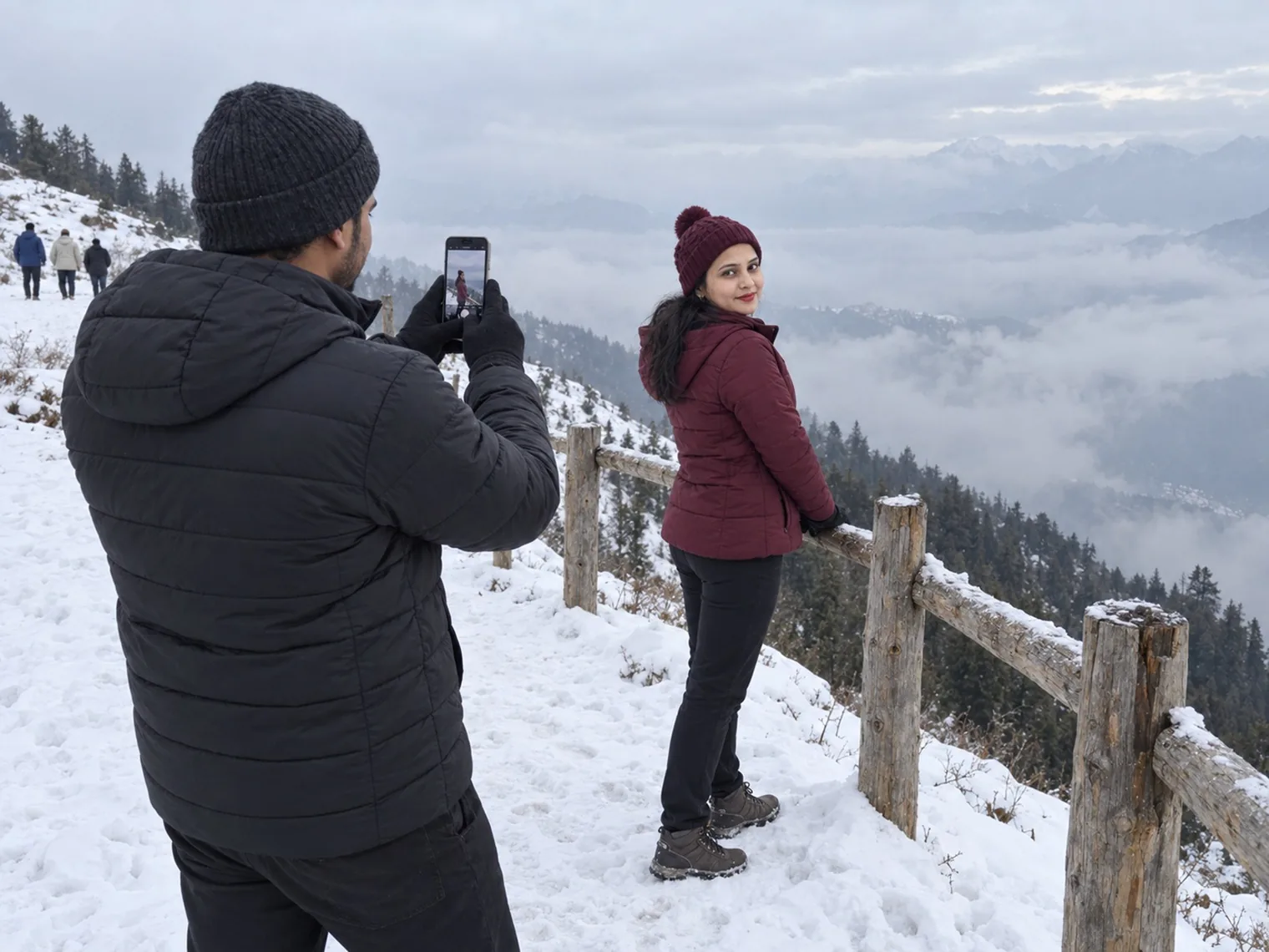 Nav photographing Vandana on his phone by a wooden fence on a snowy slope in January 2026, cloud filling the valley below.