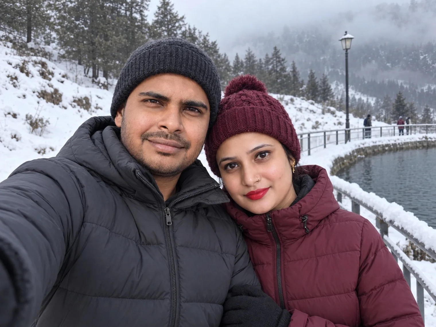 A close selfie of Nav and Vandana in winter hats in January 2026, the lake railing and snowy slope behind them.