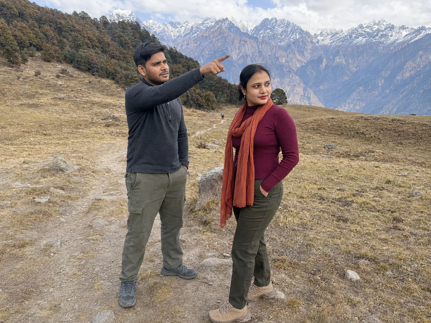 Nav pointing towards distant snow peaks while Vandana in a scarf looks the same way, the meadow around them dry and brown.