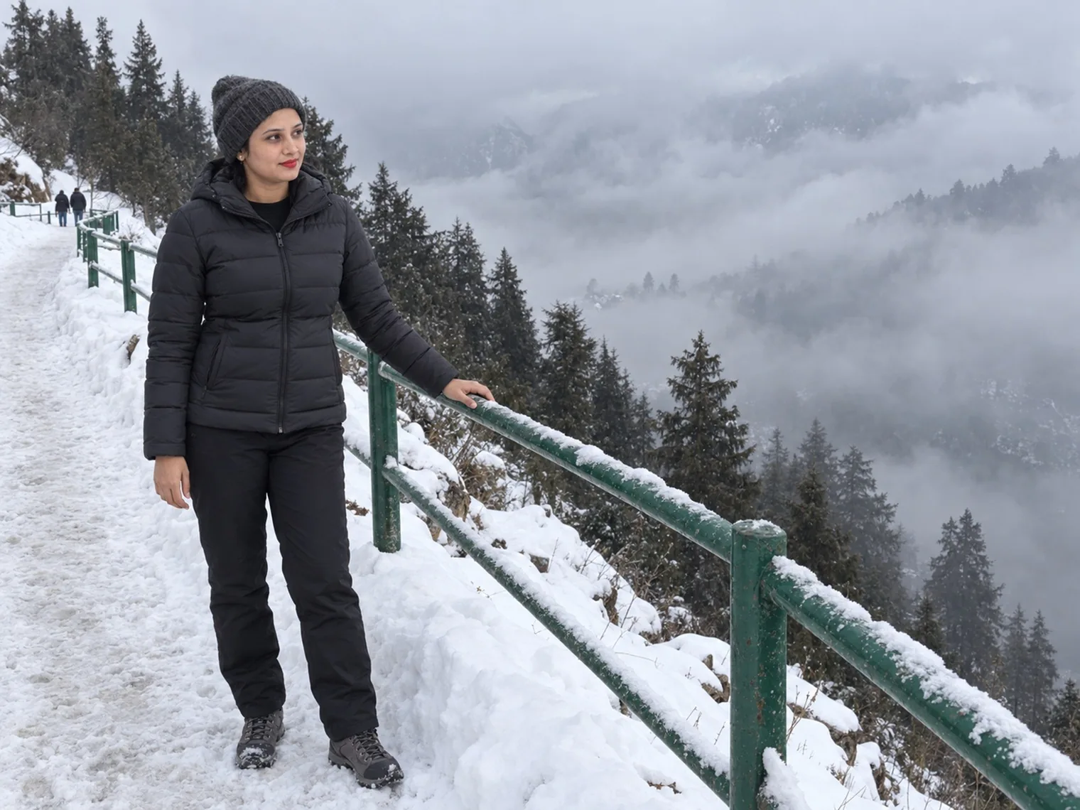 Vandana in a black puffer jacket and woollen cap on a snow-covered path beside a green railing, pine trees below her and the valley filled with cloud.