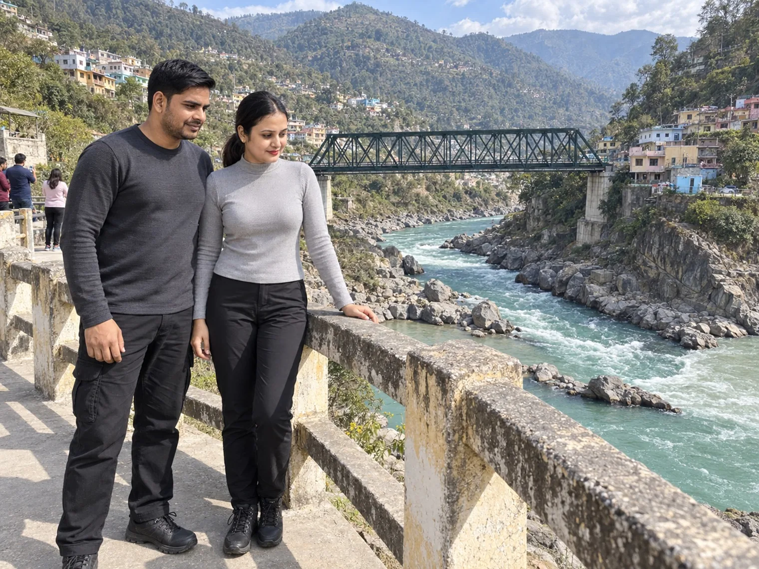Nav and Vandana standing at a concrete railing above a fast green river, a steel truss bridge crossing it and houses climbing the hillside beyond.