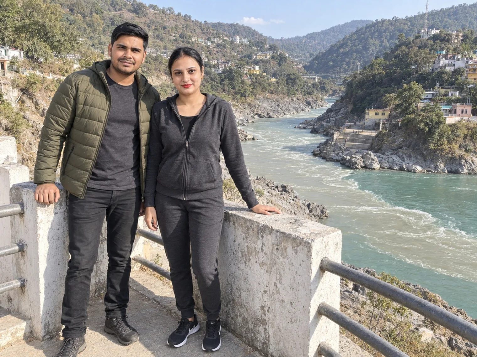 Nav and Vandana at a roadside railing above a green river where two streams meet, houses on the hillside across the water.