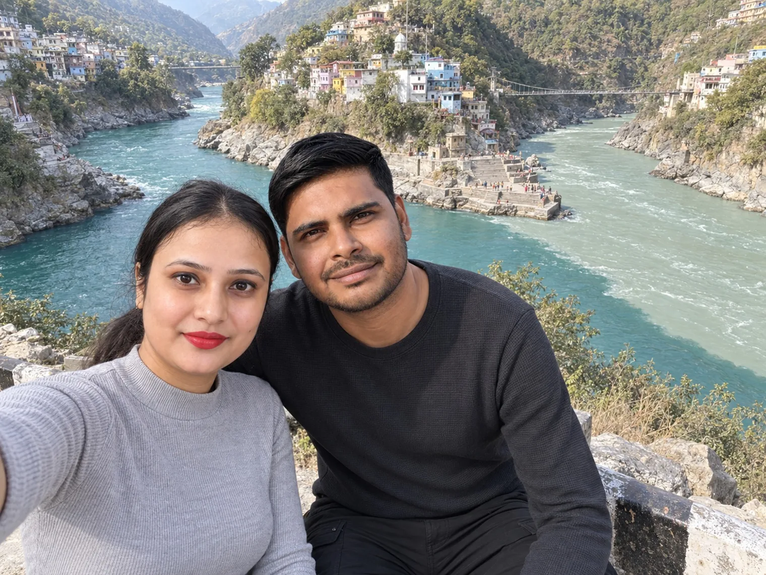 Nav and Vandana taking a selfie above a turquoise river meeting another, a hillside town and a footbridge behind.