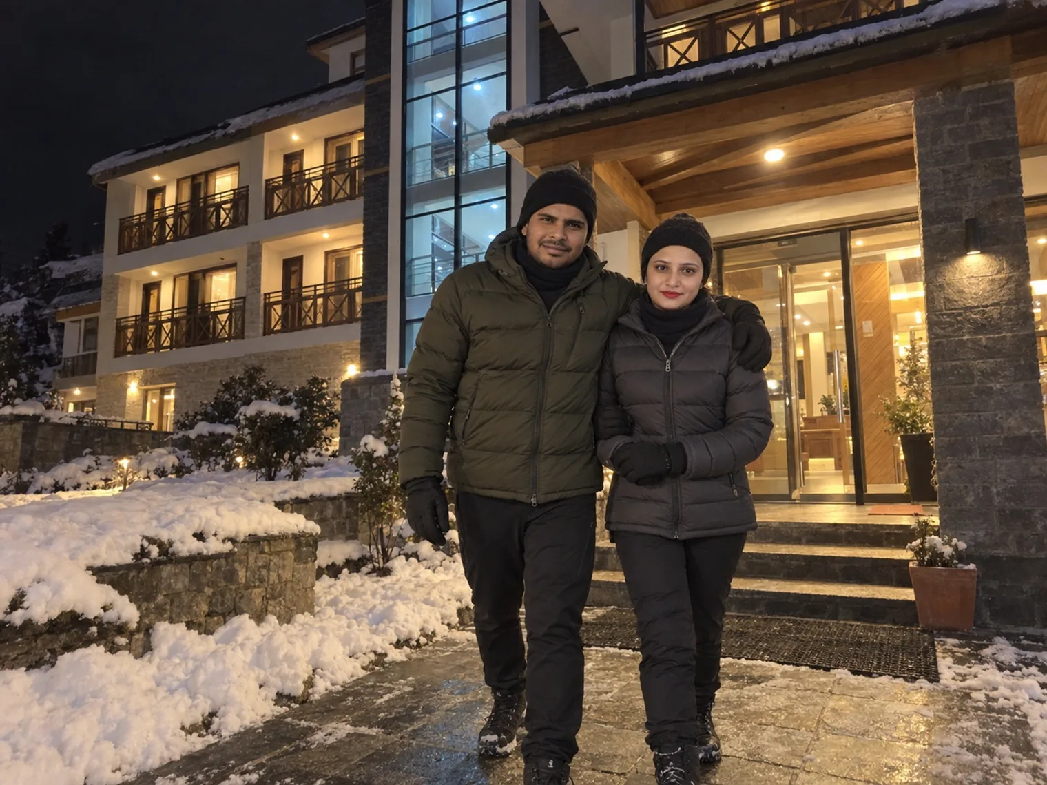 Nav and Vandana in winter jackets outside a lit hotel entrance at night, snow on the ground and a three-storey wing with balconies behind.