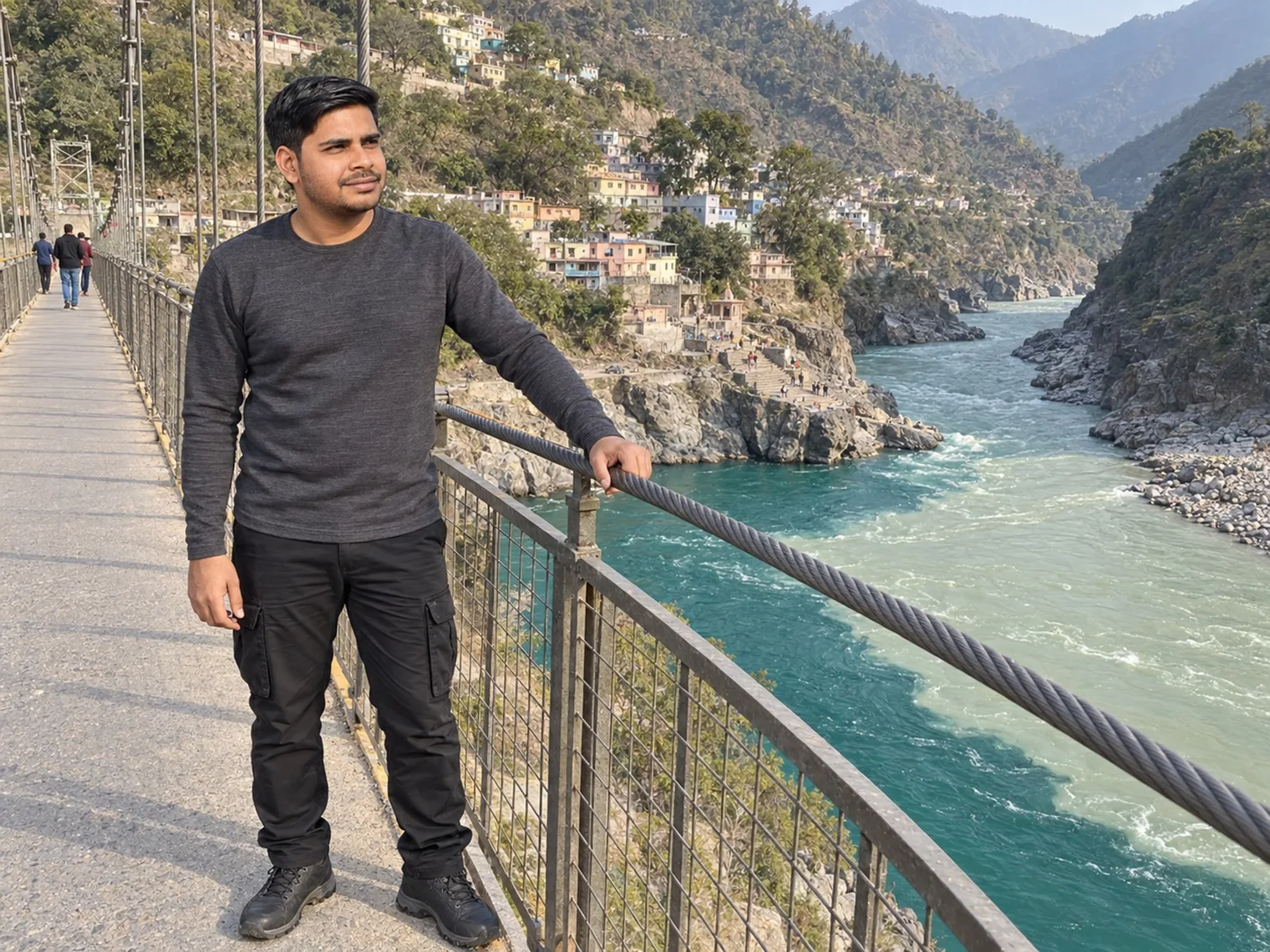 Nav standing on a steel suspension footbridge above a fast turquoise river, houses stacked on the hillside behind.