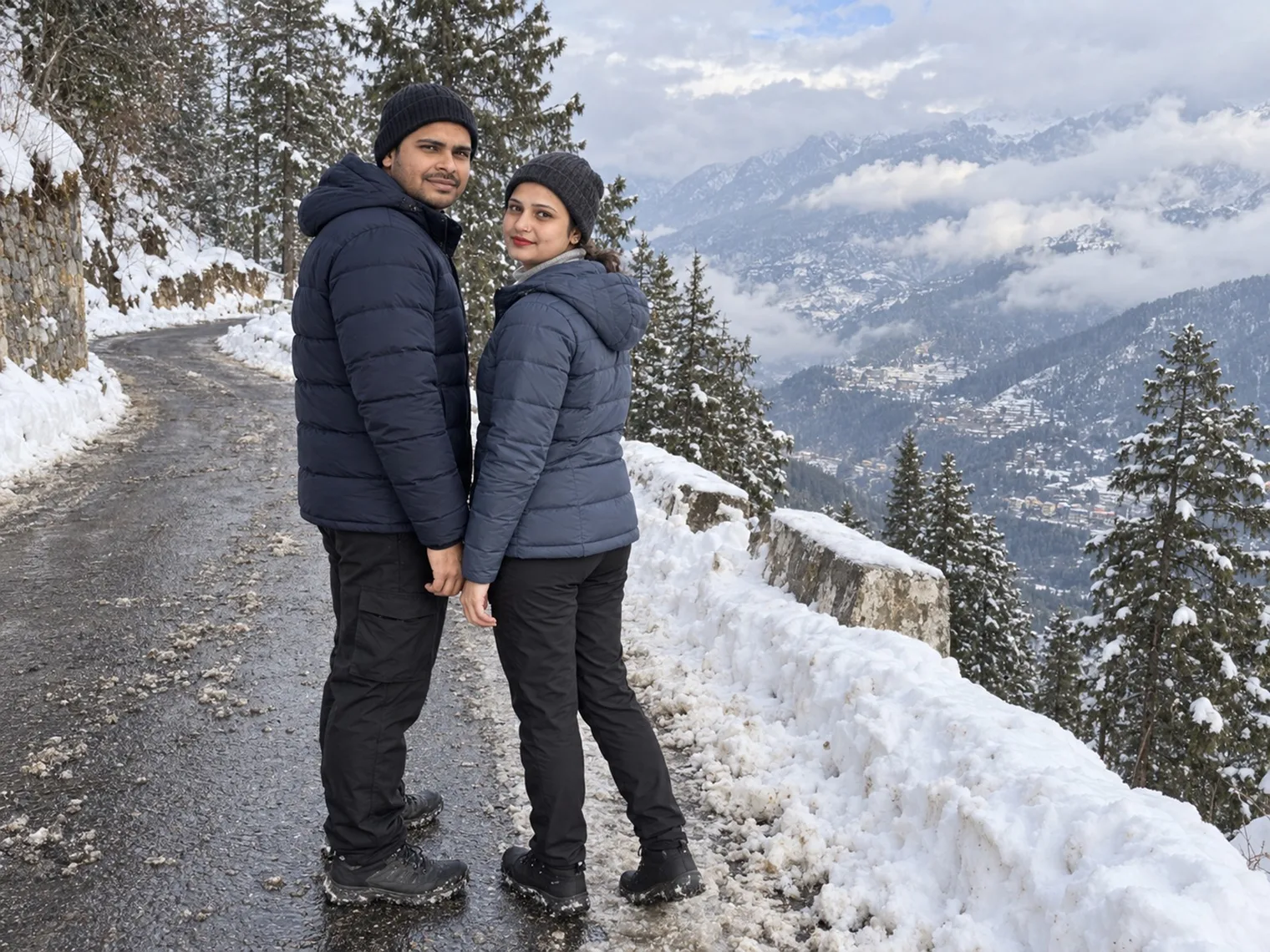 Nav and Vandana standing on a wet, slushy road cut into a snowy slope, pine trees and a valley town under cloud below them.