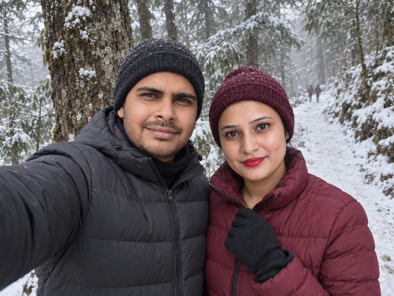 Two visitors taking a selfie in winter hats in snowy woods, a trail and tree trunks behind them.