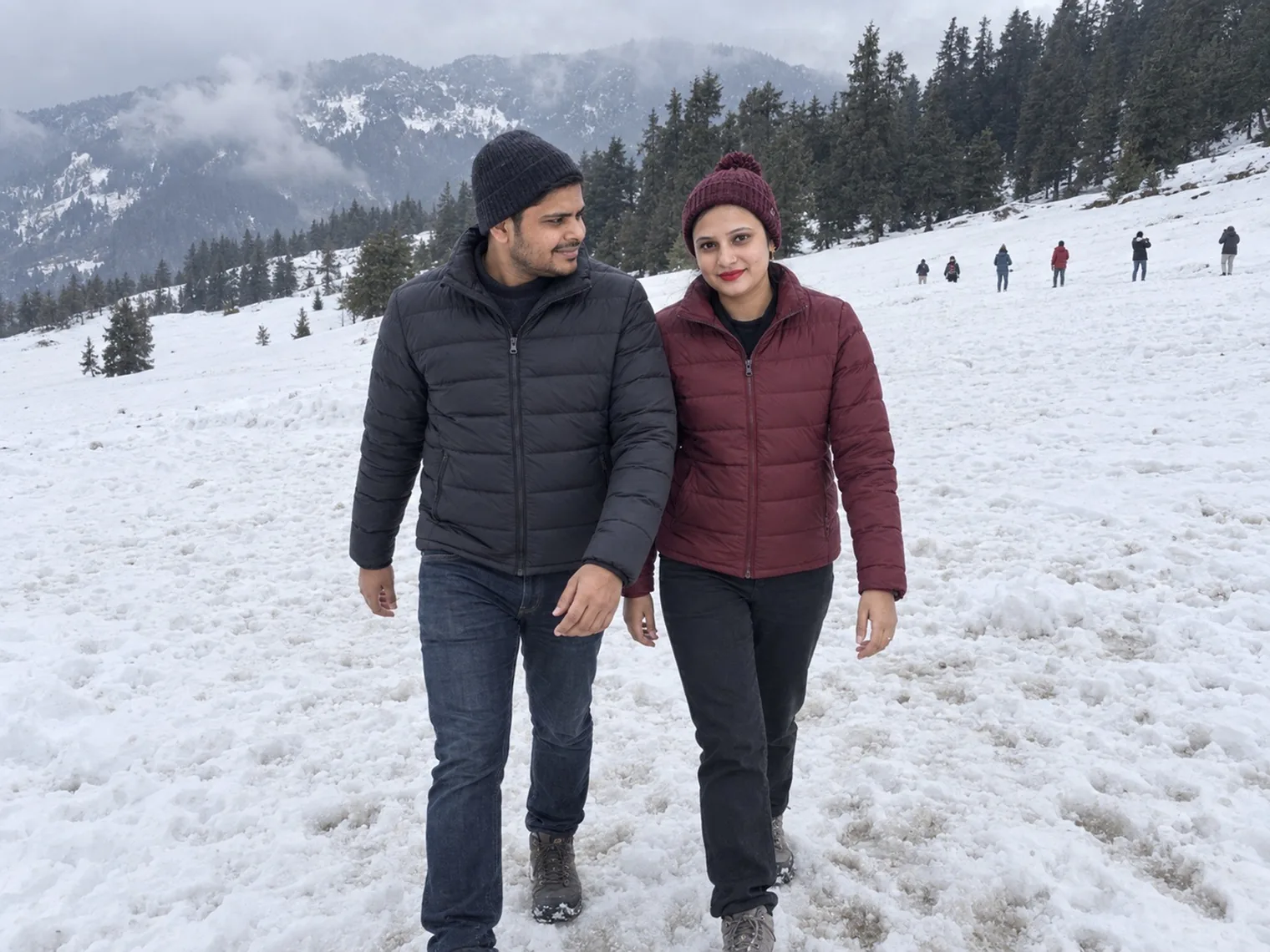 Two visitors in winter jackets walking across a snow-covered slope, more people further up the hill, pines and cloud behind.